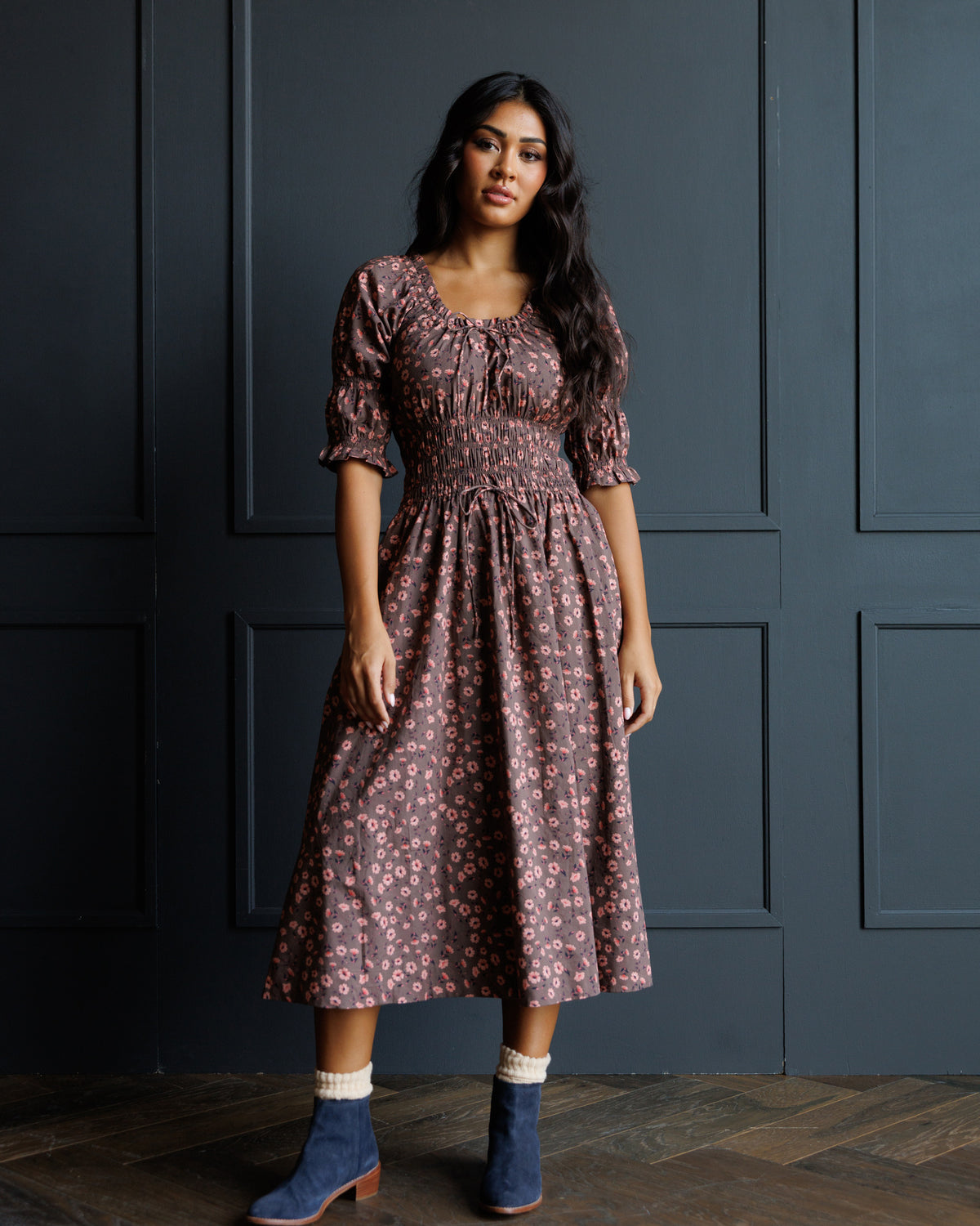 A woman stands in front of a dark paneled wall wearing a dusty rose midi dress with a small floral print, puff sleeves, and a gathered waist. She is also wearing blue ankle boots with white socks.