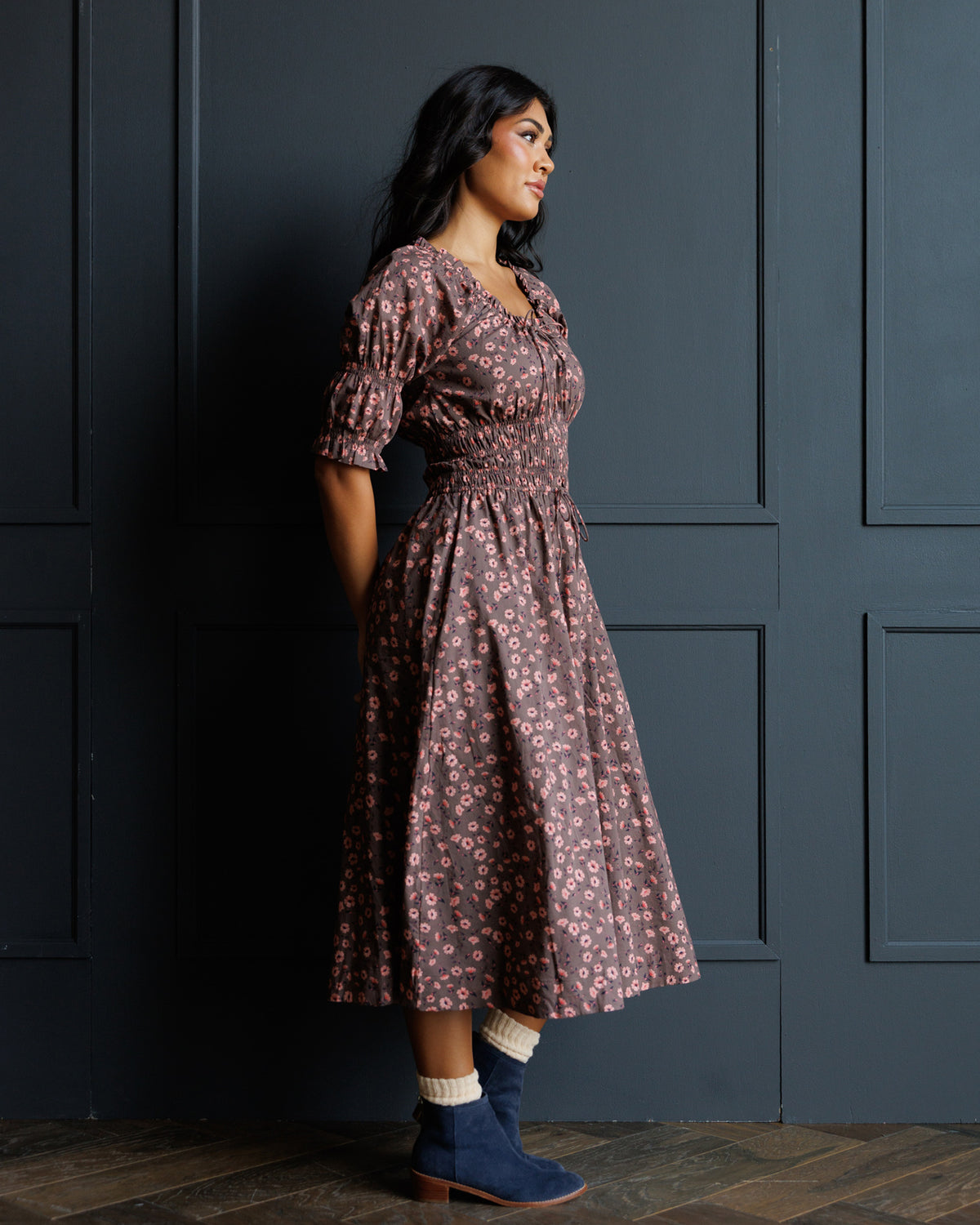 A woman wearing a brown and pink floral midi dress with puff sleeves, standing in profile against a dark paneled wall.