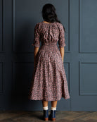 A woman viewed from behind wearing a dusty rose and brown floral print midi dress with a smocked waist and puff sleeves, standing against a dark blue paneled wall.