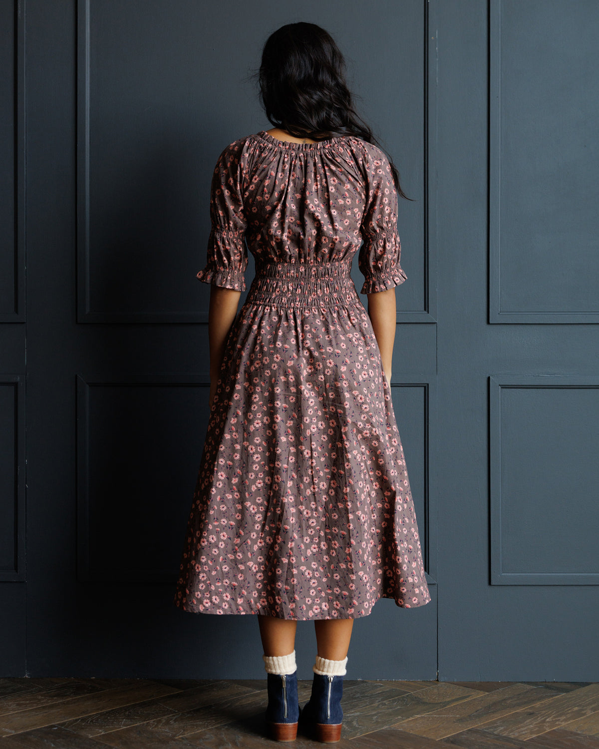 A woman viewed from behind wearing a dusty rose and brown floral print midi dress with a smocked waist and puff sleeves, standing against a dark blue paneled wall.