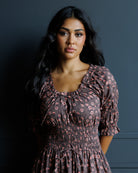 A woman with long dark hair wearing a dark brown smocked dress with a pink and white floral pattern, posing against a dark gray wall.