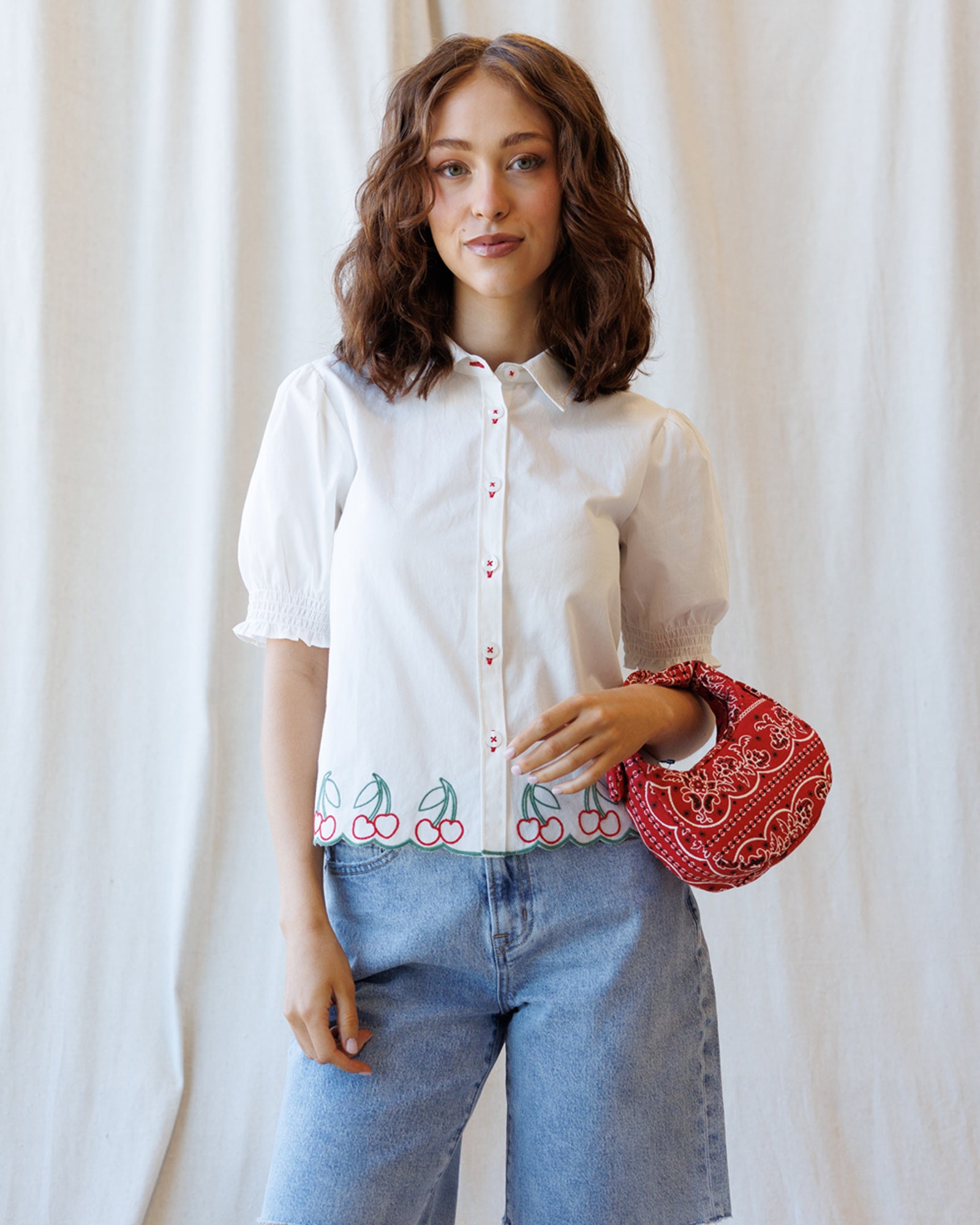 Woman wearing a white blouse with embroidery and holding a red patterned bag against a white curtain background