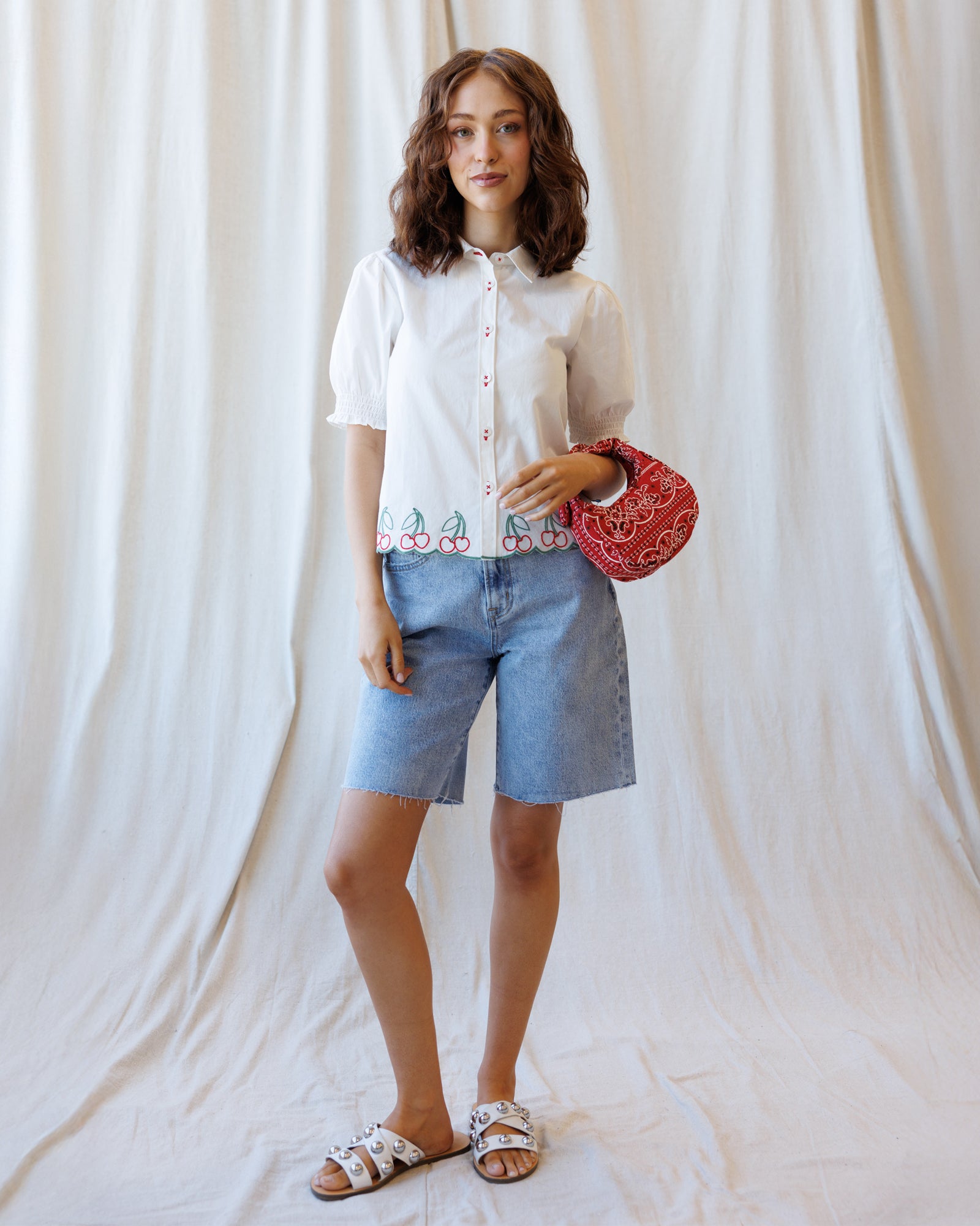 Woman wearing a white blouse with floral embroidery and denim shorts, holding a red clutch against a plain background.