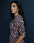 A woman with long, wavy black hair is shown from the waist up, wearing a dark brownish-gray puff-sleeve dress with a pink and white floral pattern. She is posing against a dark, paneled wall.
