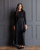 A woman modeling a long-sleeve black maxi dress with a fitted bodice and a pleated skirt, standing against a dark gray paneled wall on a wood floor.