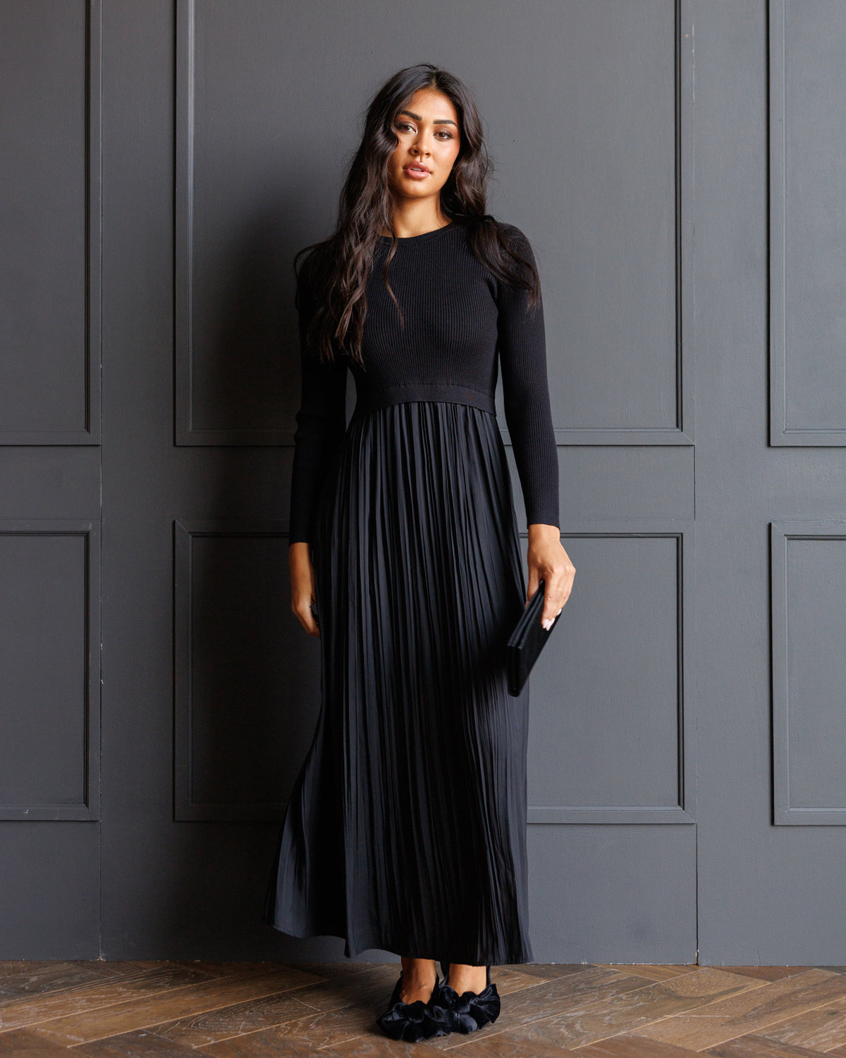 A woman modeling a long-sleeve black maxi dress with a fitted bodice and a pleated skirt, standing against a dark gray paneled wall on a wood floor.