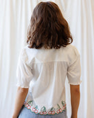 Person wearing a white blouse with cherry embroidery on a plain background