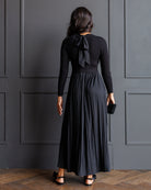A woman viewed from behind wearing a long-sleeve black top with a large bow at the back of the neck and a matching pleated maxi skirt, standing against a dark paneled wall on a wood floor.
