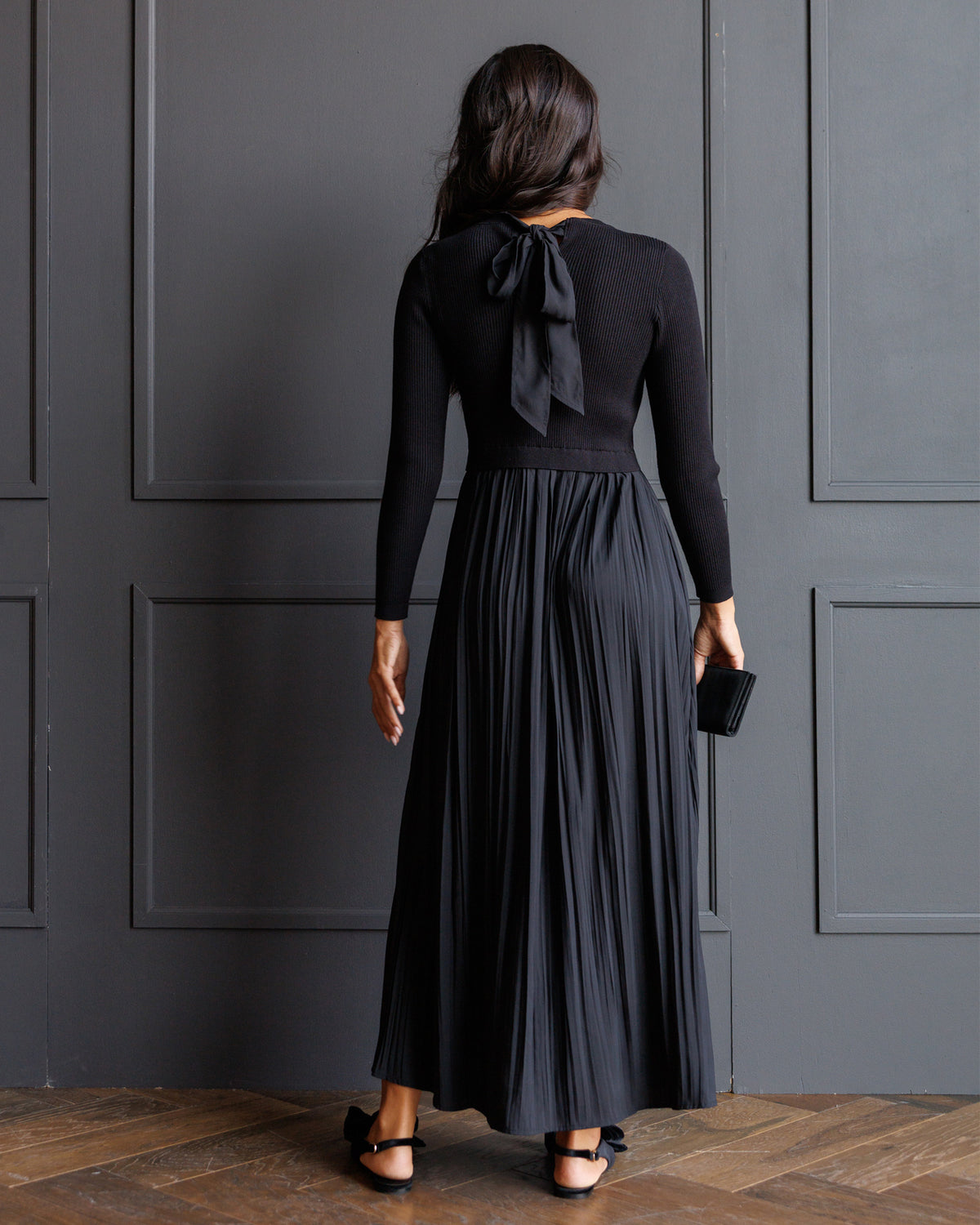 A woman viewed from behind wearing a long-sleeve black top with a large bow at the back of the neck and a matching pleated maxi skirt, standing against a dark paneled wall on a wood floor.