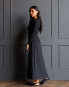 A woman wearing a black long-sleeve top and a dark charcoal pleated maxi skirt, standing against a dark gray paneled wall on a wooden floor.