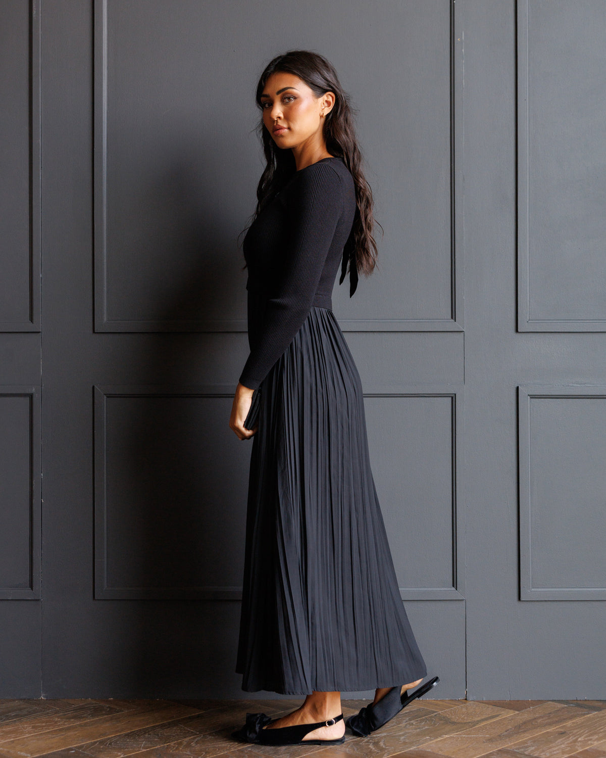 A woman wearing a black long-sleeve top and a dark charcoal pleated maxi skirt, standing against a dark gray paneled wall on a wooden floor.