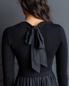 A woman viewed from the back wearing a black ribbed long-sleeve top with a large bow detail at the back neckline, paired with a matching black pleated skirt against a dark gray background.