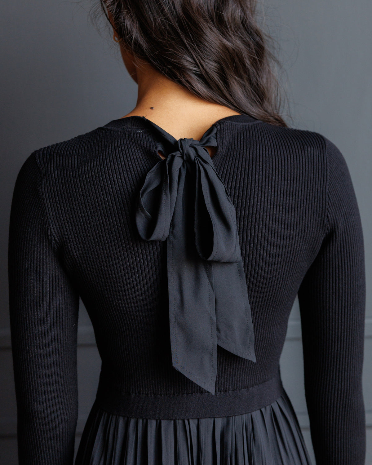 A woman viewed from the back wearing a black ribbed long-sleeve top with a large bow detail at the back neckline, paired with a matching black pleated skirt against a dark gray background.