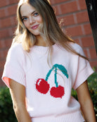 Woman wearing a white sweater with cherry design against a brick wall.