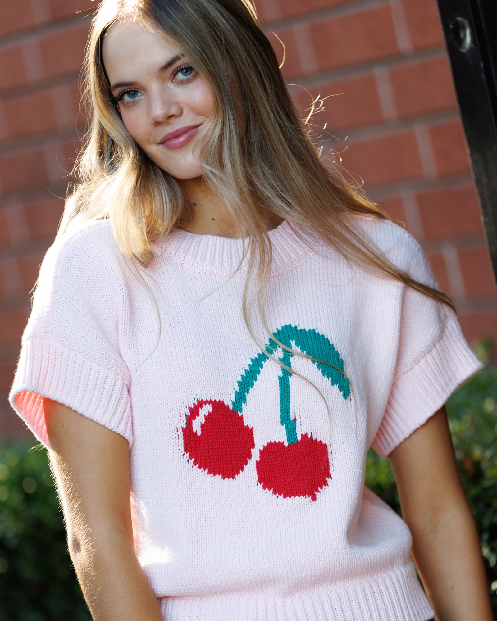 Woman wearing a white sweater with cherry design against a brick wall.