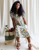 Woman in a colorful dress sitting next to a woven basket with flowers against a white curtain background