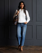 A woman stands against a dark paneled wall wearing a white long-sleeve button-up blouse with vertical ruffle details, blue jeans, and leopard-print heels, while carrying a plaid shoulder bag.