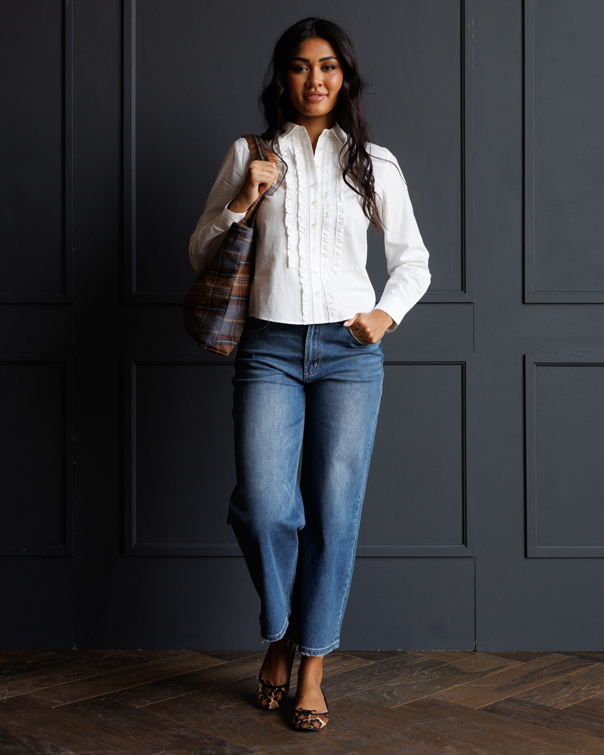A woman stands against a dark paneled wall wearing a white long-sleeve button-up blouse with vertical ruffle details, blue jeans, and leopard-print heels, while carrying a plaid shoulder bag.