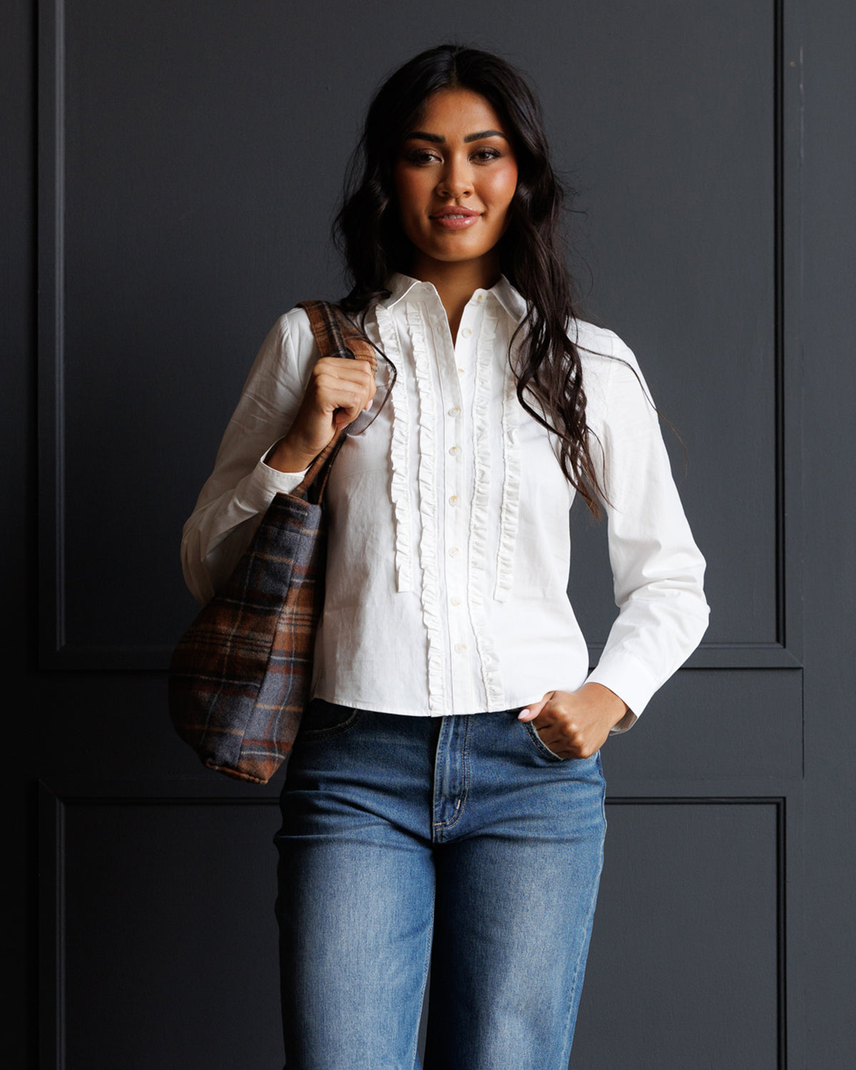 A woman modeling a white long-sleeve blouse with ruffle details down the front, paired with blue jeans and carrying a brown plaid shoulder bag, standing against a dark gray paneled wall.