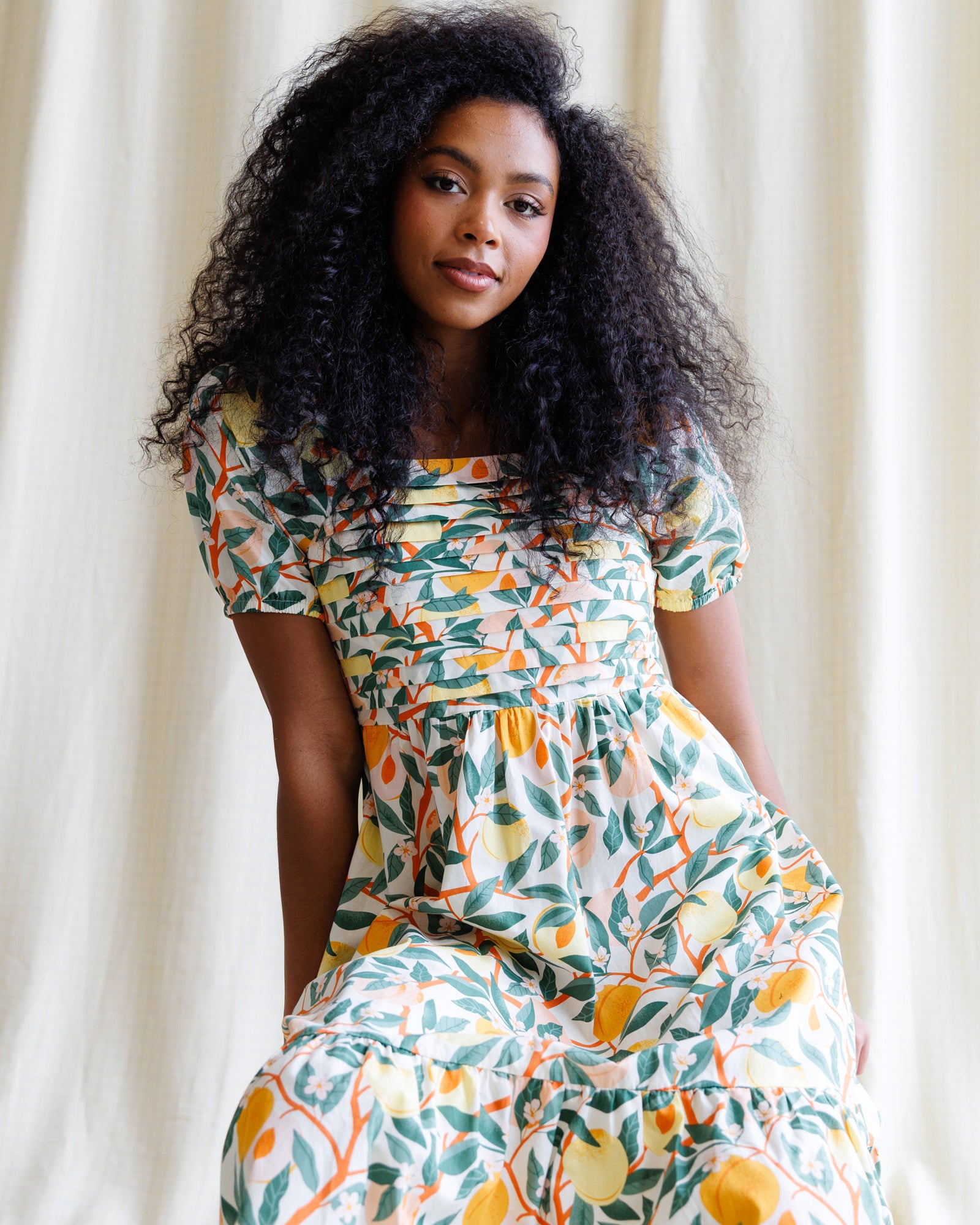 Woman wearing a colorful dress with a floral pattern against a white curtain background