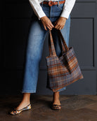 A person wearing blue jeans, a white shirt, and patterned flats holds a large plaid tote bag in brown, blue, and gray tones against a dark paneled wall.
