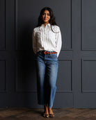 A woman stands against a dark paneled wall wearing a white long-sleeve blouse with ruffle detailing, blue jeans, a brown belt, and leopard-print flat shoes.