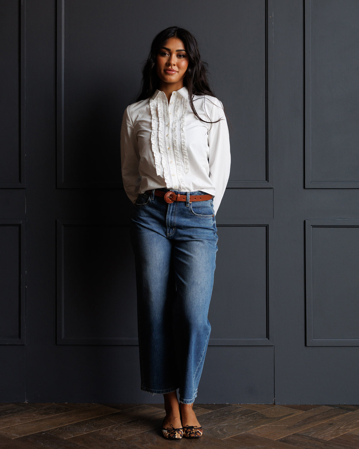 A woman stands against a dark paneled wall wearing a white long-sleeve blouse with ruffle detailing, blue jeans, a brown belt, and leopard-print flat shoes.