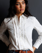 A woman wearing a white long-sleeve button-up blouse with vertical ruffle detailing down the front, paired with blue jeans and a brown belt, posed against a dark gray background.