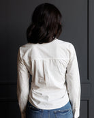 A woman viewed from behind wearing a white long-sleeve button-up shirt and blue jeans, standing against a dark gray paneled wall.