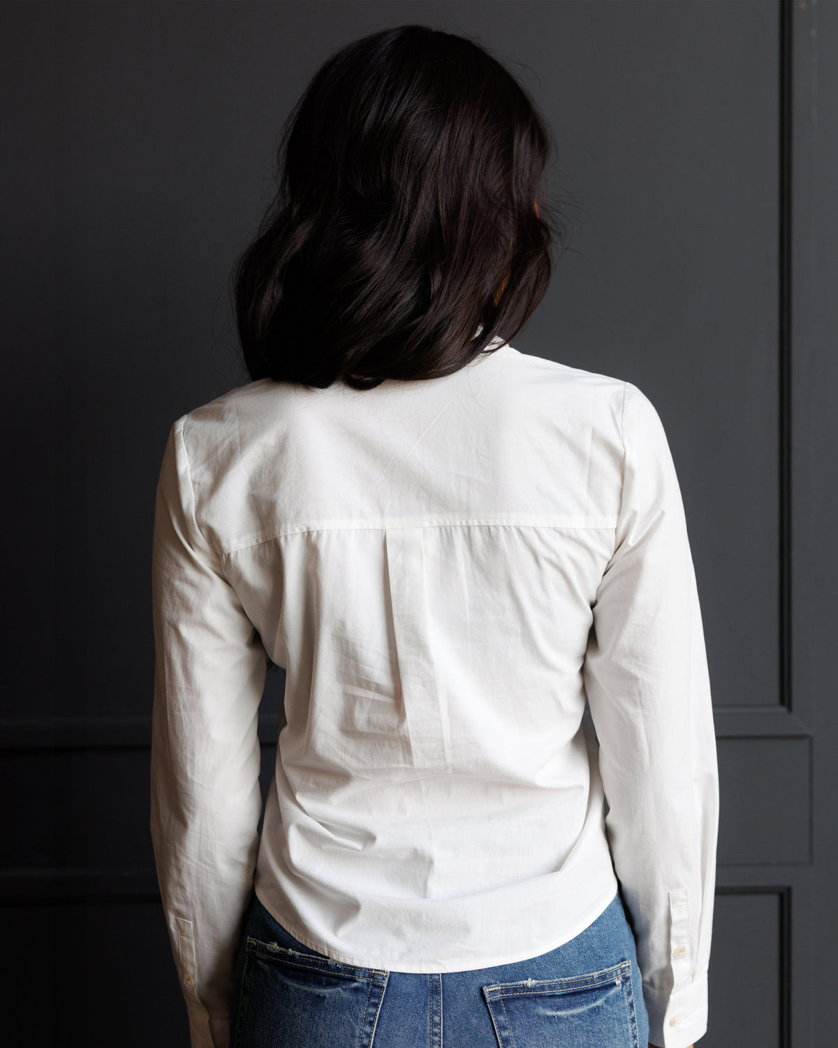 A woman viewed from behind wearing a white long-sleeve button-up shirt and blue jeans, standing against a dark gray paneled wall.
