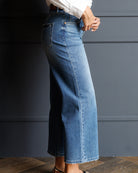A person wearing a high-waisted, wide-leg blue denim skirt with a straight front seam, paired with a white top and brown heels, standing against a dark gray paneled wall.