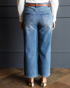 A person shown from the back wearing wide-leg cropped blue denim jeans with a brown leather belt, a white long-sleeve top, and patterned heels against a dark gray paneled wall and wood floor.