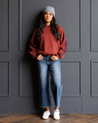 A woman stands against a dark gray paneled wall wearing a rust-red crewneck sweatshirt with blue stripes on the sleeves, blue jeans, a gray beanie, and white sneakers.