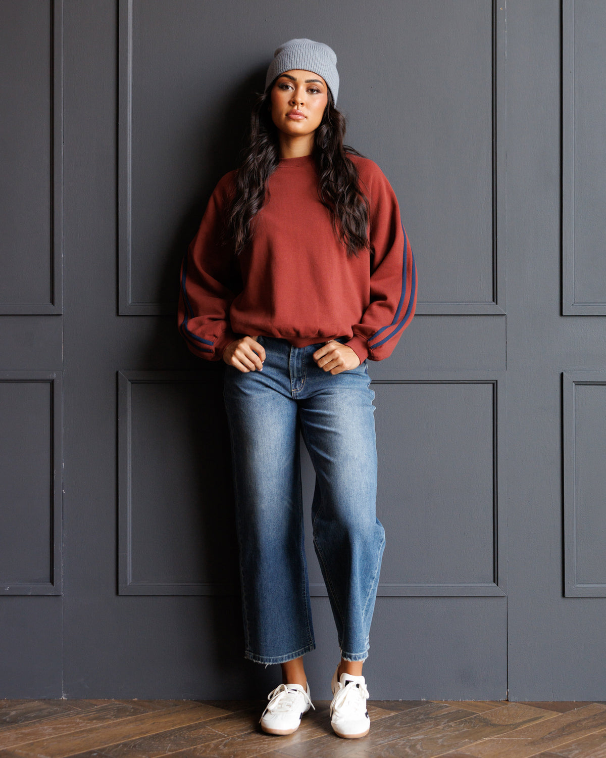 A woman stands against a dark gray paneled wall wearing a rust-red crewneck sweatshirt with blue stripes on the sleeves, blue jeans, a gray beanie, and white sneakers.