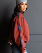 A woman wearing a rust-colored crewneck sweatshirt with blue and navy stripes down the sleeves, paired with a gray beanie and jeans, posing against a dark gray wall.