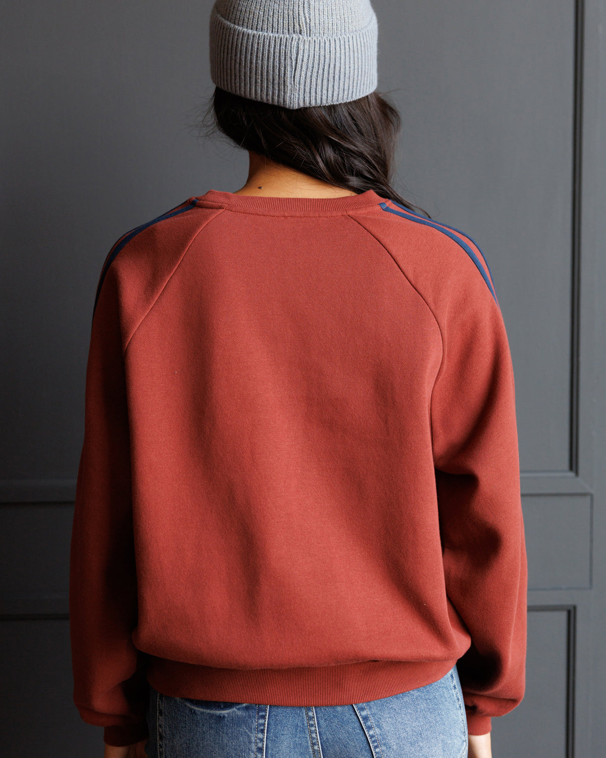 A person shown from the back wearing a rust-colored crewneck sweatshirt with blue and red stripes on the shoulders, paired with blue jeans and a light gray beanie, set against a dark gray paneled wall.