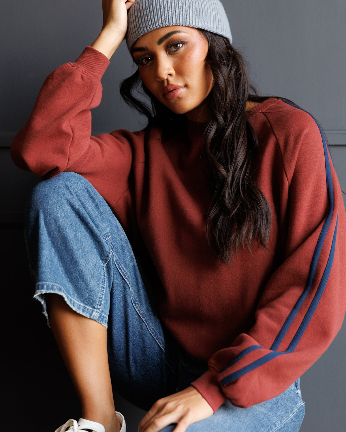 A woman wearing a rust-colored crewneck sweatshirt with blue stripes on the sleeves, blue jeans, a gray beanie, and white sneakers, posing against a dark gray wall.