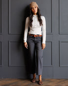 A woman stands against a dark gray paneled wall wearing a beige knit beanie, a white long-sleeve button-front top with thin dark horizontal stripes, dark gray cropped jeans, a brown belt, and leopard-print flat shoes.