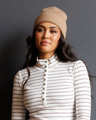 A woman wearing a beige ribbed knit beanie and a cream long-sleeve turtleneck top with thin brown horizontal stripes and a button placket, posed against a dark gray studio background.