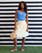 Woman in blue top and white skirt holding a straw bag against a black and white striped background