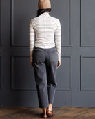 A woman is shown from behind wearing a cream and dark gray striped long-sleeve turtleneck, dark gray cropped jeans, a tan beanie, and brown woven shoes, standing in front of a dark gray paneled wall.
