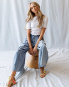 Woman sitting on a wooden stool wearing a white top and blue pants against a plain background