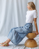 Woman sitting on a wooden stool wearing a white top and blue denim pants against a plain background