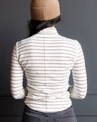 A person shown from the back wearing a fitted long-sleeve top with thin horizontal brown stripes on an off-white base, paired with dark gray jeans and a tan knit beanie against a dark gray wall.