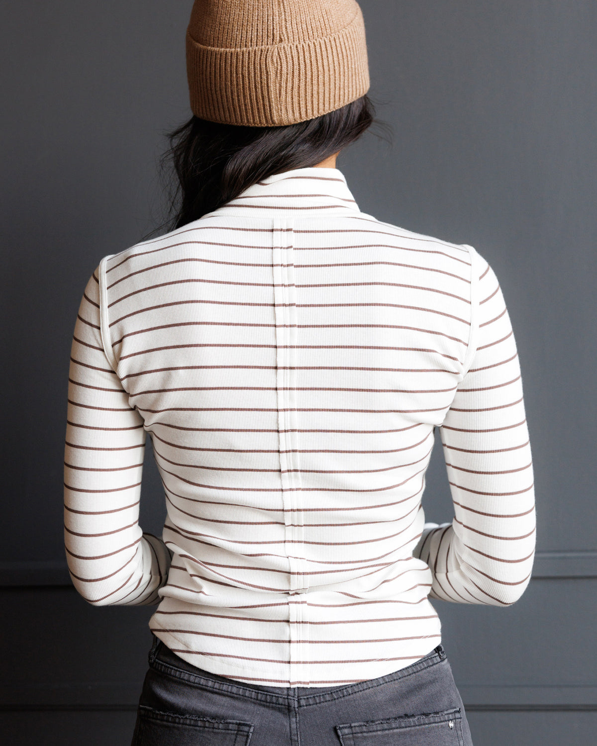 A person shown from the back wearing a fitted long-sleeve top with thin horizontal brown stripes on an off-white base, paired with dark gray jeans and a tan knit beanie against a dark gray wall.