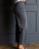 A person shown from the waist down wearing dark charcoal gray wide-leg cropped jeans, styled with a striped top, an orange-brown belt, and patterned flat shoes against a dark paneled wall.