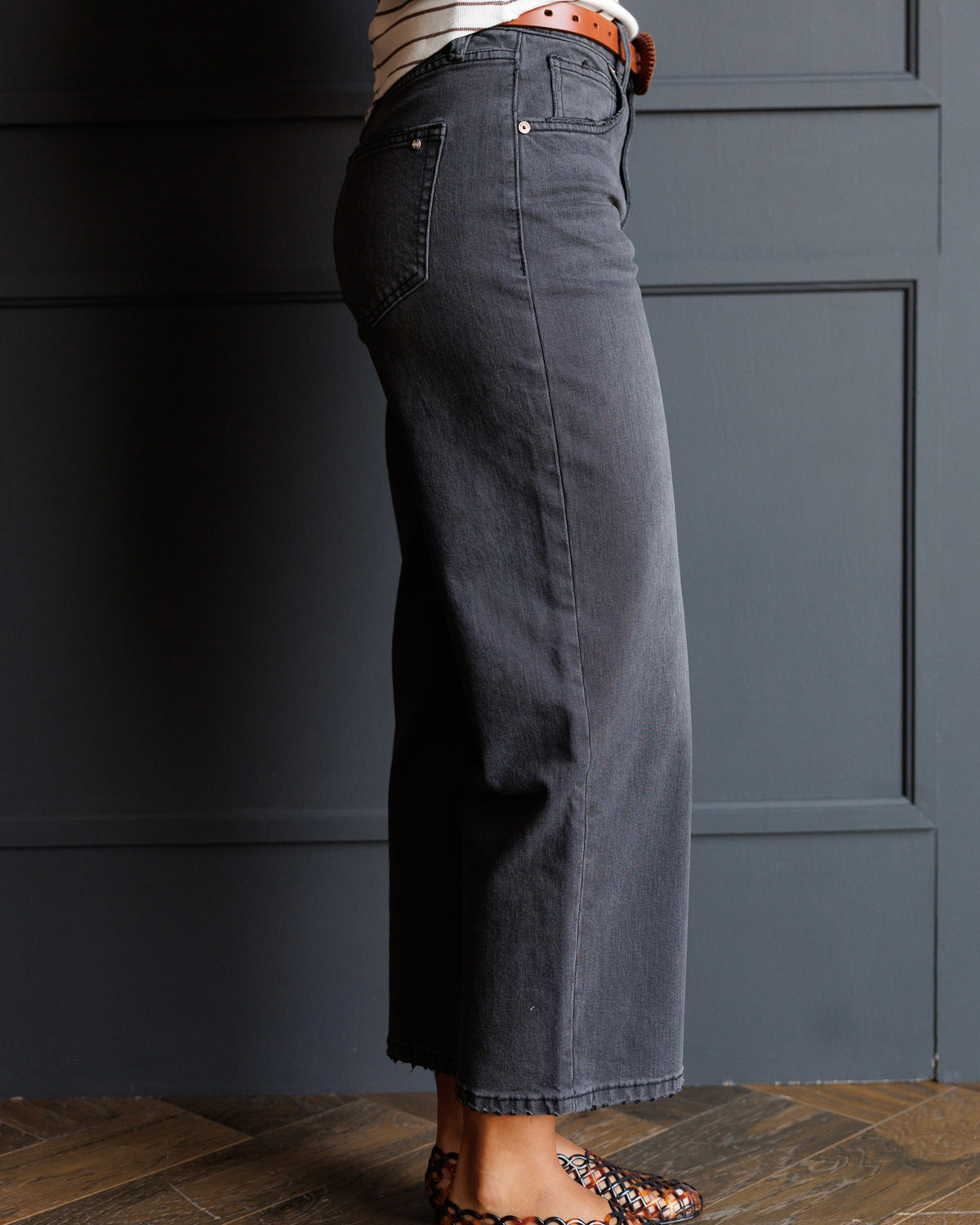 A person shown from the waist down wearing dark charcoal gray wide-leg cropped jeans, styled with a striped top, an orange-brown belt, and patterned flat shoes against a dark paneled wall.