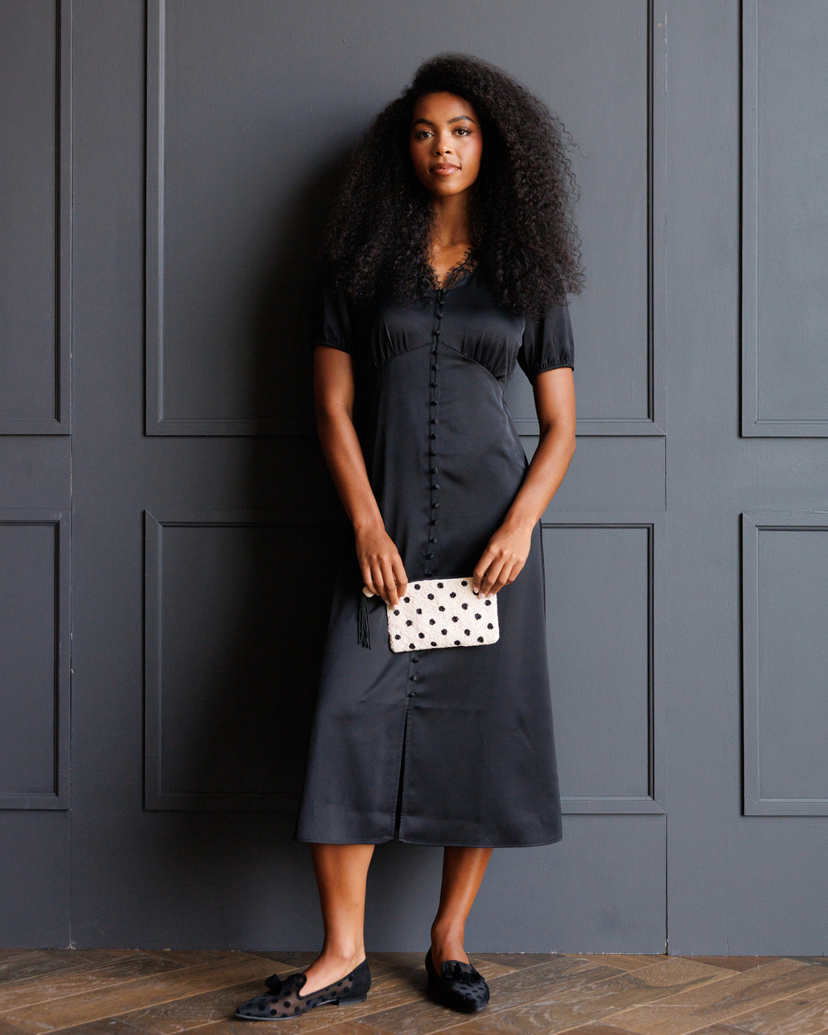 A woman with voluminous dark curly hair stands against a dark gray paneled wall wearing a short-sleeved black midi dress with a gathered front and button details. She is holding a small white clutch with black polka dots and wearing black patterned flats.