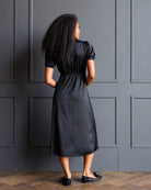 A woman with long, curly black hair is shown from behind wearing a black midi dress with short puff sleeves and black flats, standing against a dark gray paneled wall.