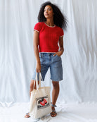 Woman in a red top and denim shorts holding a tote bag against a white background